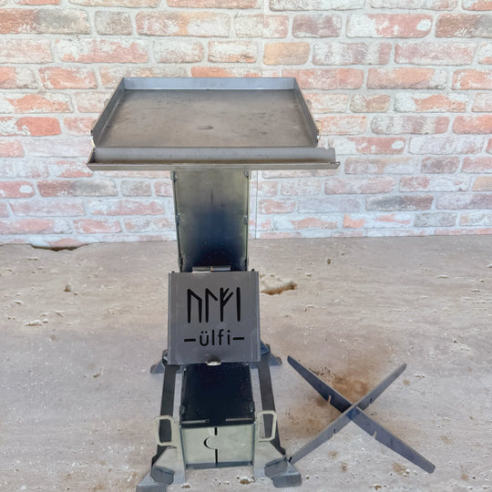 Brasero en métal avec plateau de cuisson sur socle robuste devant un mur de briques.