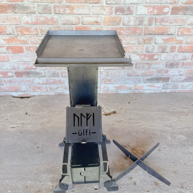 Brasero en métal avec plateau de cuisson sur socle robuste devant un mur de briques.