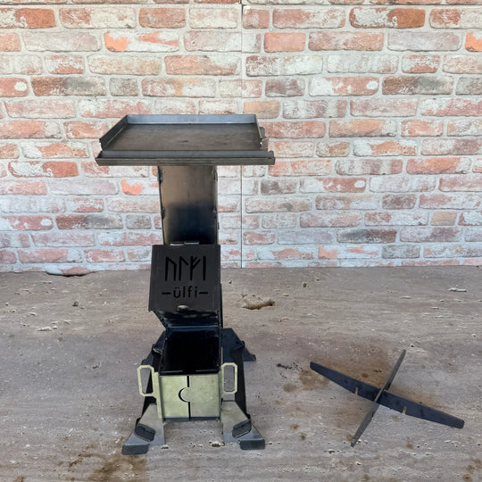 Brasero en métal noir design sur socle avec support pour bois, devant un mur de briques.